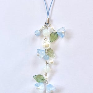 May include: A delicate blue and white floral charm with a light blue cord. The charm features small blue flower beads, white pearl beads, and green leaf accents, all connected by silver-toned metal. A charming accessory.