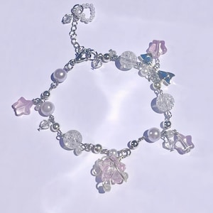 May include: A delicate bracelet featuring a silver chain adorned with a variety of beads. The bracelet includes clear, iridescent, and pearl-like beads, along with pink star-shaped charms and a cluster of pink beads. A small ring of pearls is attached.