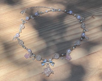 May include: A delicate necklace featuring a silver chain adorned with faux pearls, clear crystal beads, and small pink star-shaped charms. A silver bow and a pink crystal pendant hang from the center. The necklace is displayed on a wooden surface.