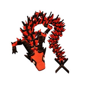 May include: A red and black articulated dragon toy with a detailed design. The dragon has a red body with black spikes and accents. The head features white eyes and black horns. The tail ends in a black cross shape.