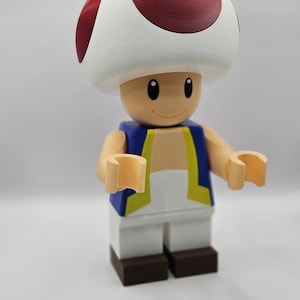 May include: A large, colorful figurine of the character Toad from the Super Mario video game series. The figure has a white mushroom cap with red spots, a tan face, and a blue vest with yellow trim. The figure is made of plastic.