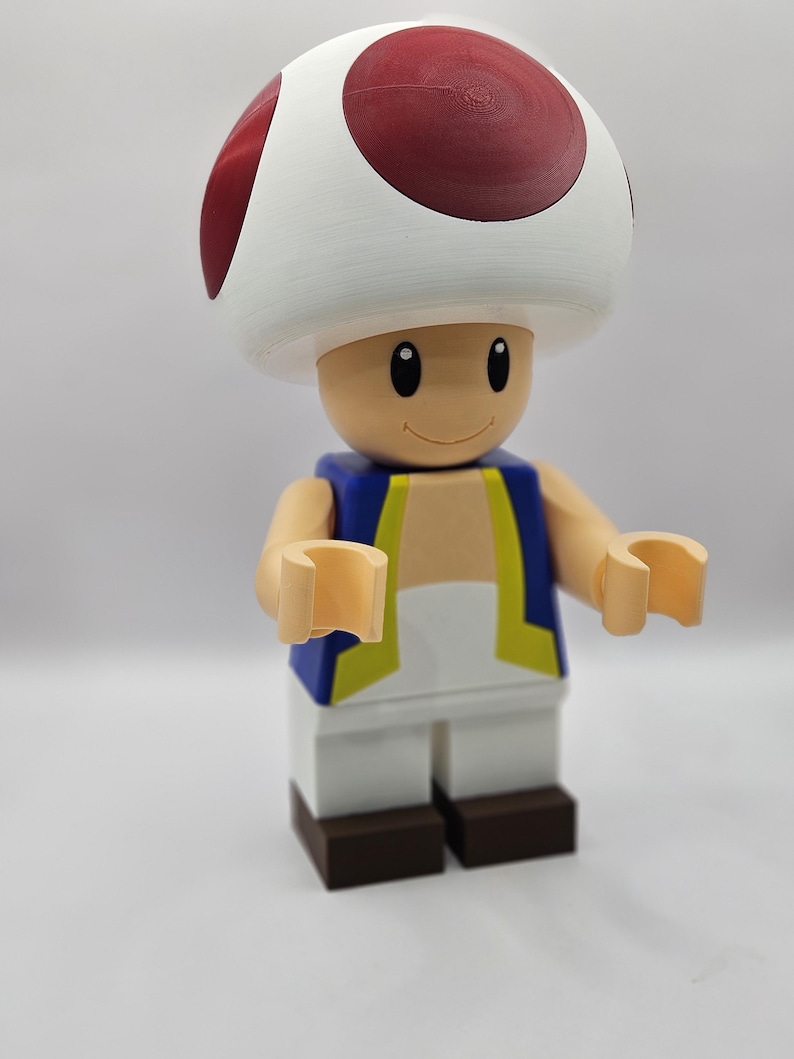 May include: A large, colorful figurine of the character Toad from the Super Mario video game series. The figure has a white mushroom cap with red spots, a tan face, and a blue vest with yellow trim. The figure is made of plastic.