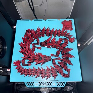 May include: A red 3D-printed dragon model, disassembled on a light blue surface. The dragon is composed of multiple spiky segments, with a detailed head and body. The model is in a 3D printer, with tools visible below.