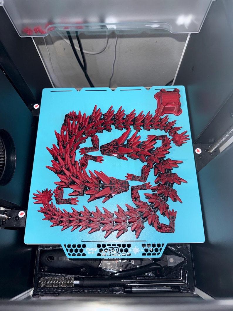 May include: A red 3D-printed dragon model, disassembled on a light blue surface. The dragon is composed of multiple spiky segments, with a detailed head and body. The model is in a 3D printer, with tools visible below.