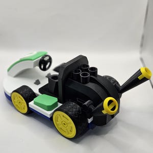 May include: A 3D-printed toy race car in black, white, green, and yellow. The car features a black body with yellow accents, a white base, and green and blue details. The wheels are yellow with black tires, and it includes a green and white remote control.