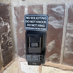 May include: A black Ring doorbell camera with two white and black signs attached. The top sign reads "NO SOLICITING DO NOT KNOCK DO NOT RING WORKING FROM HOME." The bottom sign says "DO NOT DISTURB" and "ring."