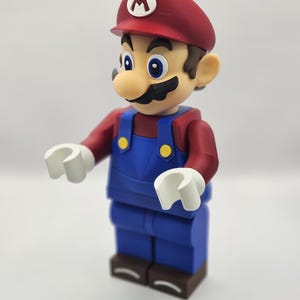Op de afbeelding: Een beeldje van het videogamepersonage Mario, met een rode pet met een witte "M", een rood shirt, een blauwe overall en witte handen. De figuur heeft een grote neus en een zwarte snor. De figuur staat op een bruine basis.