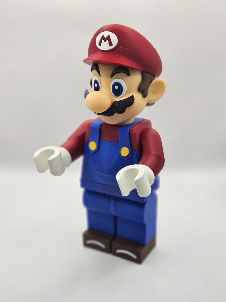 Op de afbeelding: Een beeldje van het videogamepersonage Mario, met een rode pet met een witte "M", een rood shirt, een blauwe overall en witte handen. De figuur heeft een grote neus en een zwarte snor. De figuur staat op een bruine basis.