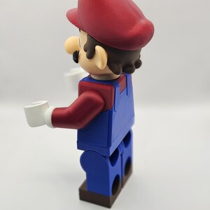 Op de afbeelding: Een 3D-geprint figuur van Mario, het iconische videogamepersonage. Het figuur is in blokstijl, met een rode hoed, blauwe overall en witte handschoenen. Het figuur houdt twee witte blokken vast.