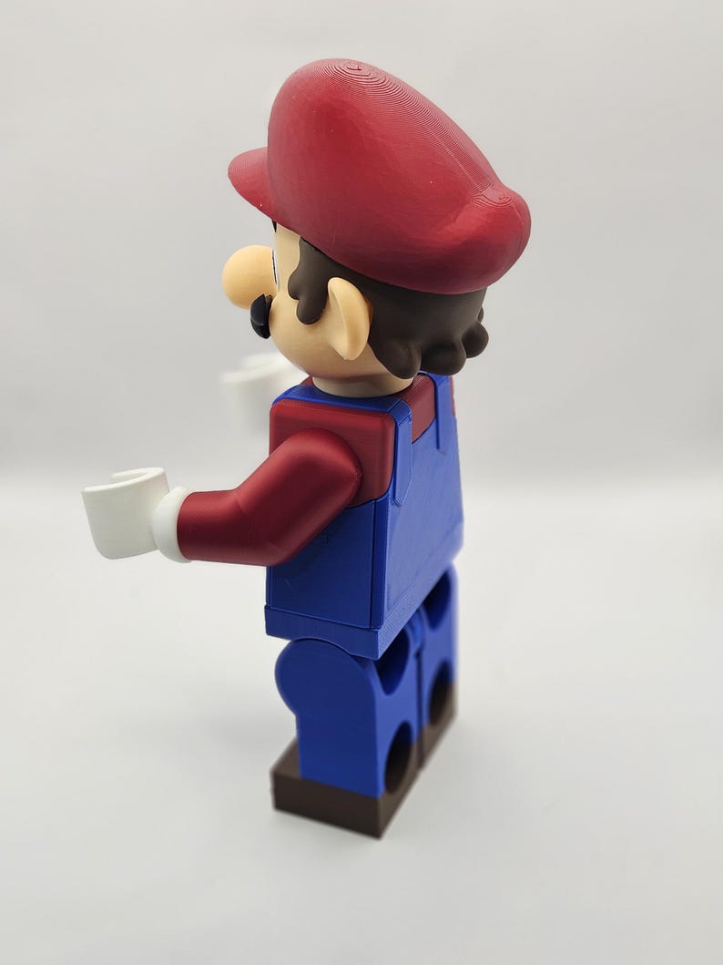Op de afbeelding: Een 3D-geprint figuur van Mario, het iconische videogamepersonage. Het figuur is in blokstijl, met een rode hoed, blauwe overall en witte handschoenen. Het figuur houdt twee witte blokken vast.