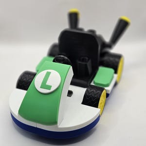 May include: A 3D-printed toy kart in green, white, black, blue, and yellow. The kart features a large green panel with a white circle and the letter "L". The tires are black with yellow rims. The kart has a black seat and steering wheel.