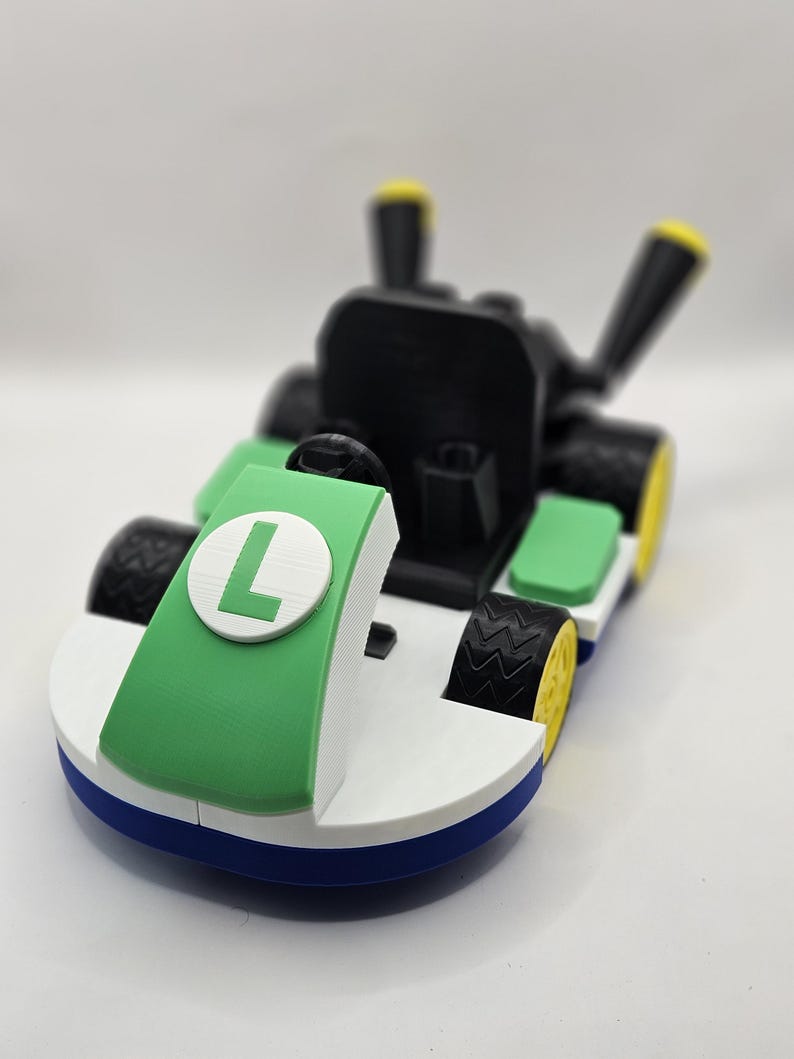 May include: A 3D-printed toy kart in green, white, black, blue, and yellow. The kart features a large green panel with a white circle and the letter "L". The tires are black with yellow rims. The kart has a black seat and steering wheel.