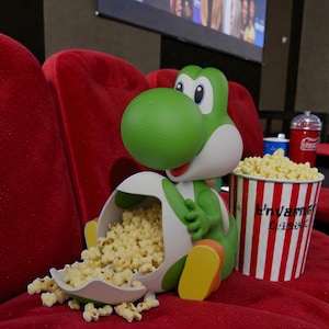 May include: A green Yoshi figurine spilling popcorn from a white container, with a red and white striped popcorn bucket. The scene is set in a movie theater with a red seat and a screen in the background.