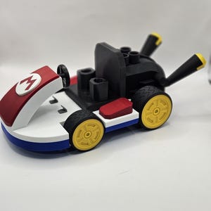 May include: A toy kart in red, white, blue, black, and yellow. The kart features a red front with a white "M" logo, a black seat, and yellow wheels. The kart is a toy.
