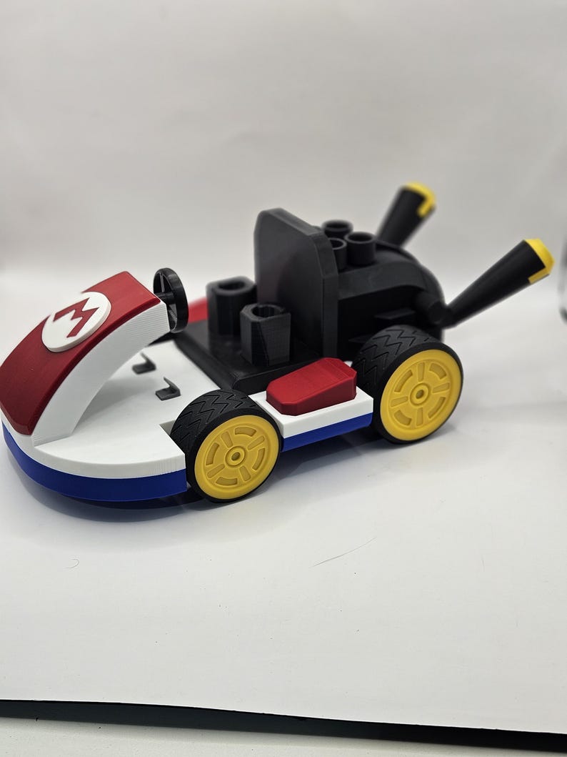 May include: A toy kart in red, white, blue, black, and yellow. The kart features a red front with a white "M" logo, a black seat, and yellow wheels. The kart is a toy.