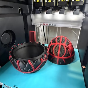 May include: A 3D printer is shown with two black and red decorative objects. One object is a bowl with a lotus flower design, and the other is a spherical object with a spider web design. The objects are on a blue surface.