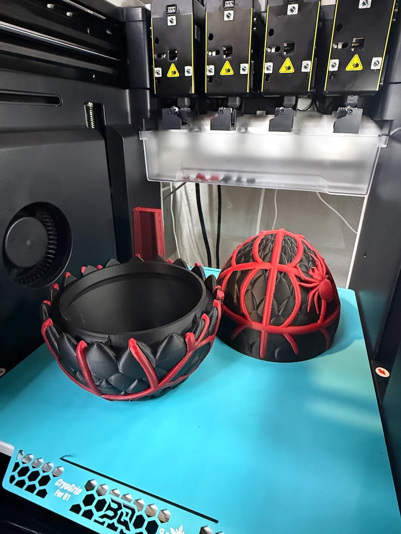 May include: A 3D printer is shown with two black and red decorative objects. One object is a bowl with a lotus flower design, and the other is a spherical object with a spider web design. The objects are on a blue surface.