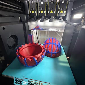 May include: A 3D printer with two red and blue decorative objects. One is a bowl-shaped item, and the other is a half-sphere. The objects are on a light blue surface. The printer has multiple black components.