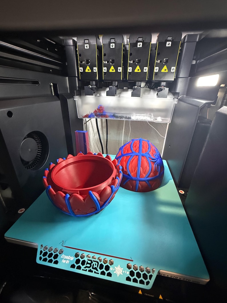 May include: A 3D printer with two red and blue decorative objects. One is a bowl-shaped item, and the other is a half-sphere. The objects are on a light blue surface. The printer has multiple black components.