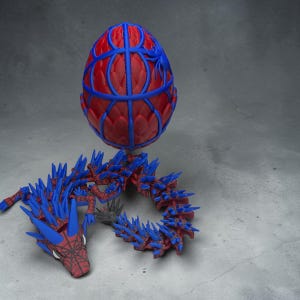 May include: A red and blue 3D-printed dragon toy with a matching egg. The dragon has blue spikes and a red body. The egg is red with blue web-like detailing. The toy is set against a gray background.