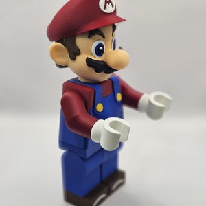 Op de afbeelding: Een beeldje van het videogamepersonage Mario. De figuur draagt een rode hoed met een witte "M" erop, een rood shirt met lange mouwen, een blauwe overall en bruine schoenen. De figuur heeft witte handen en een grote neus.