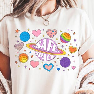 Puede incluir: Camiseta color crema con un diseño gráfico colorido. El diseño incluye un planeta con las palabras "SAFE SPACE" y varios corazones, estrellas y planetas en tonos rosa, morado, azul y naranja.