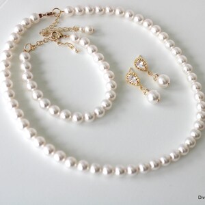 K&ouml;nnte beinhalten: Ein Schmuckset bestehend aus einer wei&szlig;en Perlenkette, einem passenden Perlenarmband und zwei Ohrh&auml;ngern aus goldfarbenem Metall mit klaren tropfenf&ouml;rmigen Steinen und kleinen Perlen. Der Text Divine Jewel ist unten rechts aufgedruckt.