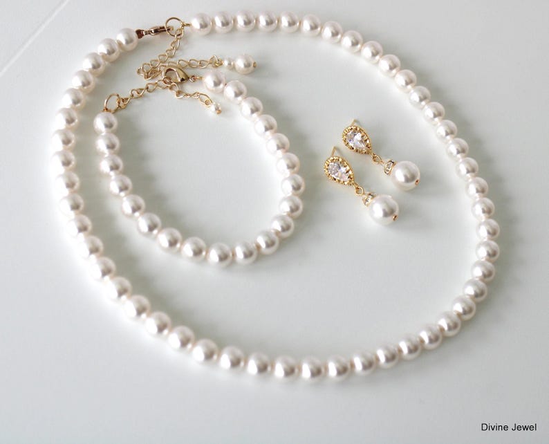 K&ouml;nnte beinhalten: Ein Schmuckset bestehend aus einer wei&szlig;en Perlenkette, einem passenden Perlenarmband und zwei Ohrh&auml;ngern aus goldfarbenem Metall mit klaren tropfenf&ouml;rmigen Steinen und kleinen Perlen. Der Text Divine Jewel ist unten rechts aufgedruckt.