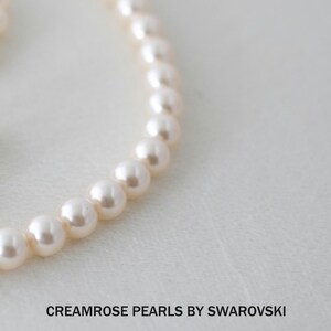 May include: A strand of cream-colored round pearls arranged in a curved line. The text CREAMROSE PEARLS BY SWAROVSKI is printed in black below the pearls, with the word Divinejewel appearing in the bottom right corner of the frame.