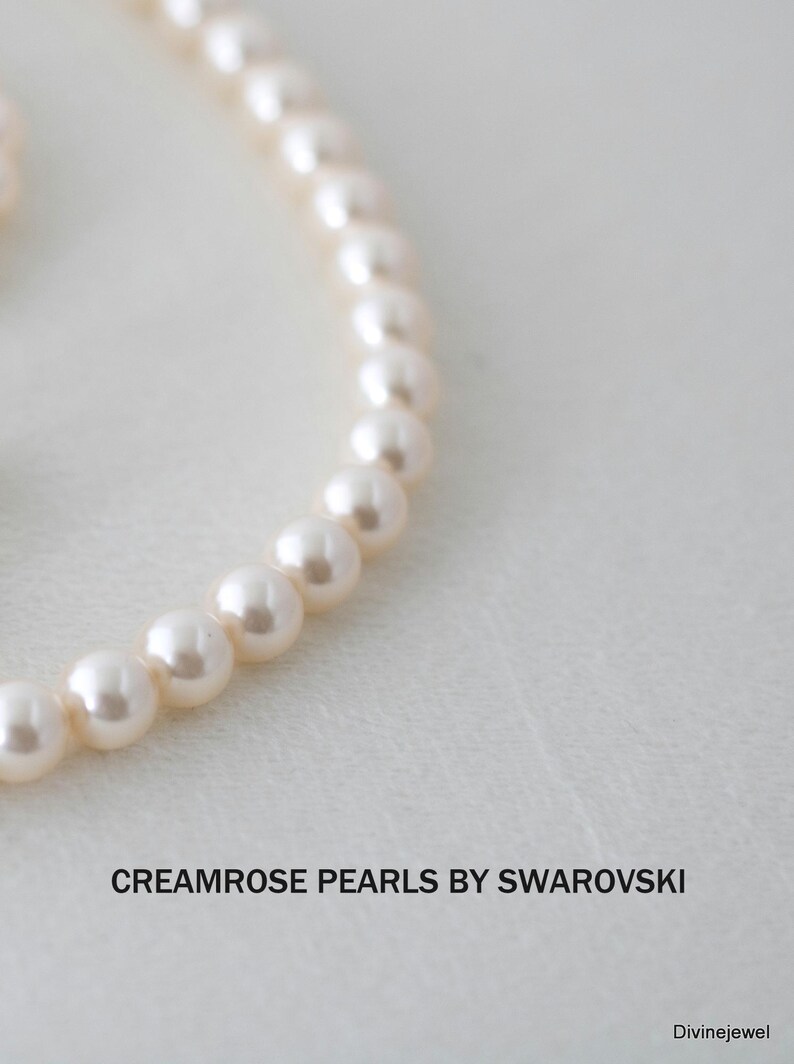May include: A strand of cream-colored round pearls arranged in a curved line. The text CREAMROSE PEARLS BY SWAROVSKI is printed in black below the pearls, with the word Divinejewel appearing in the bottom right corner of the frame.