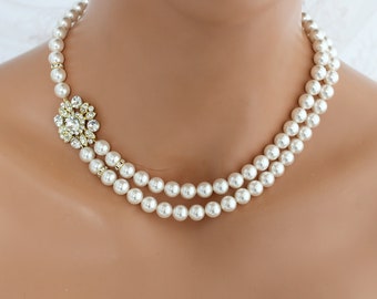 Op de afbeelding: Een parelketting met twee strengen en een goudkleurige bloemversiering. De ketting heeft ronde, crèmekleurige parels en een cluster van heldere kristallen in het midden van het bloemmotief. De ketting is ontworpen om om de nek te worden gedragen.