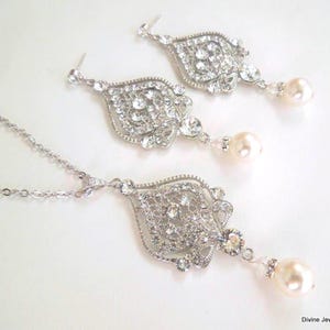 A silver-colored necklace and matching earrings, each with an ornate, filigree-style pendant encrusted with clear crystals and a single pearl drop, displayed on a white background.
