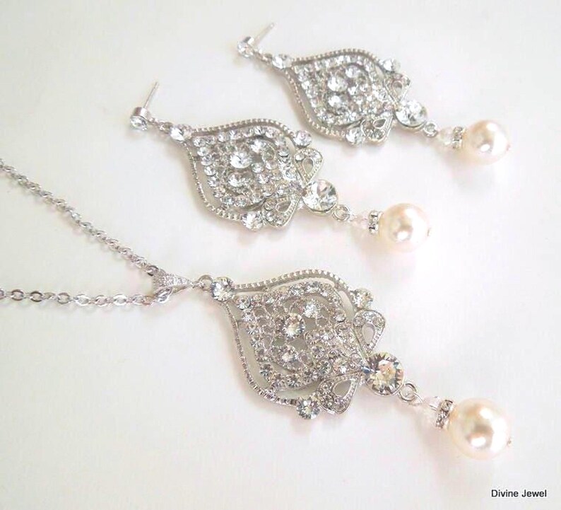 A silver-colored necklace and matching earrings, each with an ornate, filigree-style pendant encrusted with clear crystals and a single pearl drop, displayed on a white background.