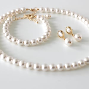 K&ouml;nnte beinhalten: Ein Schmuckset bestehend aus einer Perlenkette, einem passenden Perlenarmband und zwei Ohrh&auml;ngern. Jedes St&uuml;ck besteht aus wei&szlig;en runden Perlen und goldfarbenen Metallakzenten. Die Ohrringe enthalten einen klaren tropfenf&ouml;rmigen Stein. Text: Divine Jewel.