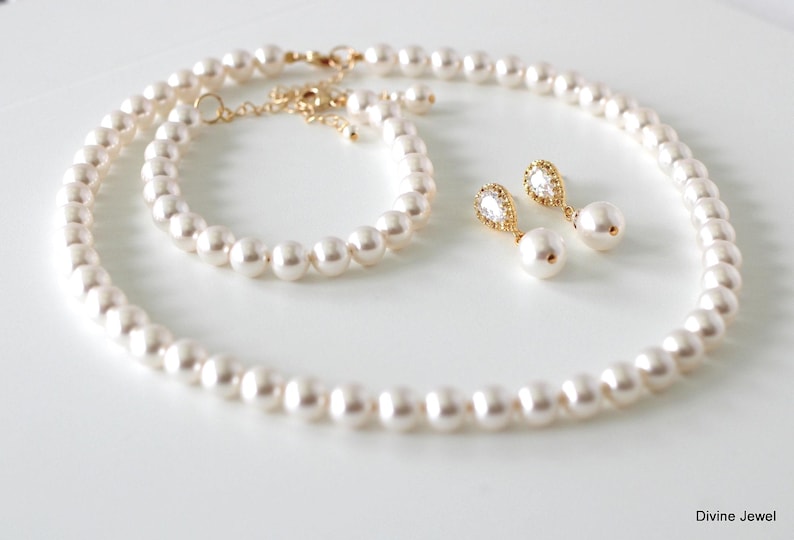 K&ouml;nnte beinhalten: Ein Schmuckset bestehend aus einer Perlenkette, einem passenden Perlenarmband und zwei Ohrh&auml;ngern. Jedes St&uuml;ck besteht aus wei&szlig;en runden Perlen und goldfarbenen Metallakzenten. Die Ohrringe enthalten einen klaren tropfenf&ouml;rmigen Stein. Text: Divine Jewel.