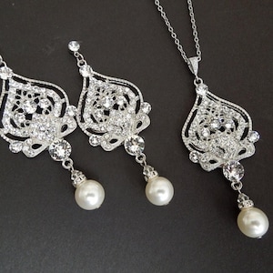 A silver-colored necklace and matching earrings, each with an ornate, filigree-style pendant encrusted with clear crystals and a single pearl drop, displayed on a dark background.