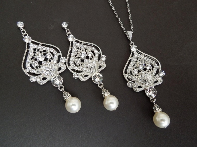 A silver-colored necklace and matching earrings, each with an ornate, filigree-style pendant encrusted with clear crystals and a single pearl drop, displayed on a dark background.