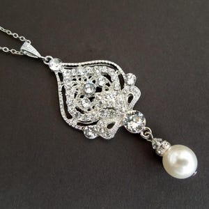 A silver-colored necklace with an ornate, filigree-style pendant encrusted with clear crystals and a single pearl drop, displayed on a dark background.