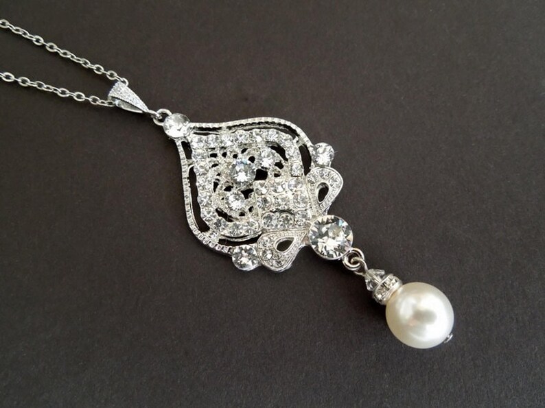 A silver-colored necklace with an ornate, filigree-style pendant encrusted with clear crystals and a single pearl drop, displayed on a dark background.