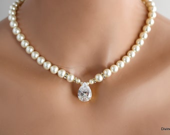 Op de afbeelding: Een eenvoudige parelketting met een grote, traanvormige kristallen hanger. De ketting is gemaakt van ivoorwitte parels en heeft een gouden sluiting.