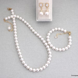 K&ouml;nnte beinhalten: Ein Schmuckset bestehend aus einer wei&szlig;en Perlenkette, einem passenden Perlenarmband und einem Paar goldfarbenen Ohrh&auml;ngern mit tropfenf&ouml;rmigen, klaren Steinen. Die Kette und das Armband verf&uuml;gen &uuml;ber goldfarbene Metallkettenverl&auml;ngerungen mit Verschl&uuml;ssen.