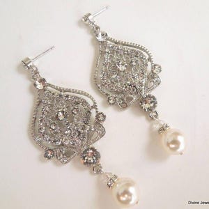 A pair of silver-colored earrings, each with an ornate, filigree-style pendant encrusted with clear crystals and a single pearl drop, displayed on a white background.