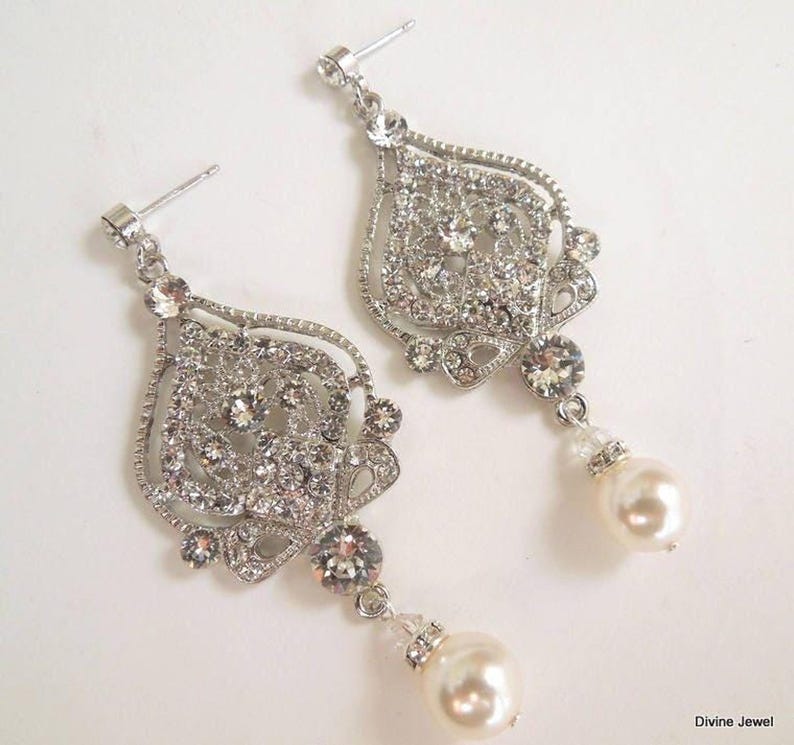 A pair of silver-colored earrings, each with an ornate, filigree-style pendant encrusted with clear crystals and a single pearl drop, displayed on a white background.