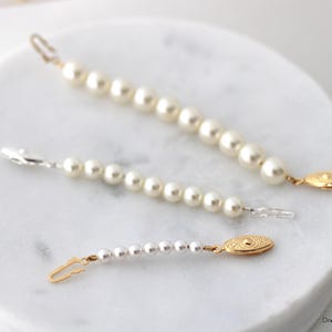 May include: Three decorative garment extenders featuring rows of white faux pearls. The top extender has a gold-tone hook and oval clasp. The middle extender has silver-tone lobster claw clasps. The bottom extender has a gold-tone hook and oval clasp.