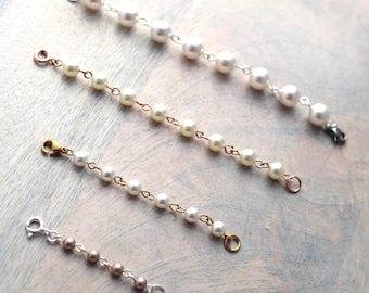 May include: Four pearl bracelet extenders of varying lengths and metal tones. The top extender is silver-toned with white pearls, followed by a rose gold-toned one, a gold-toned one, and a shorter silver-toned extender with smaller brown pearls.