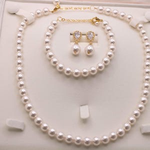 K&ouml;nnte beinhalten: Ein Schmuckset bestehend aus einer wei&szlig;en Perlenkette, einem passenden Perlenarmband und zwei Ohrh&auml;ngern. Jeder Ohrh&auml;nger besteht aus einer tropfenf&ouml;rmigen wei&szlig;en Perle, die an einer goldfarbenen Fassung mit einem klaren Stein h&auml;ngt.
