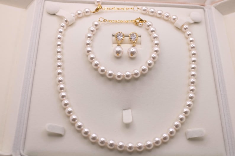 K&ouml;nnte beinhalten: Ein Schmuckset bestehend aus einer wei&szlig;en Perlenkette, einem passenden Perlenarmband und zwei Ohrh&auml;ngern. Jeder Ohrh&auml;nger besteht aus einer tropfenf&ouml;rmigen wei&szlig;en Perle, die an einer goldfarbenen Fassung mit einem klaren Stein h&auml;ngt.