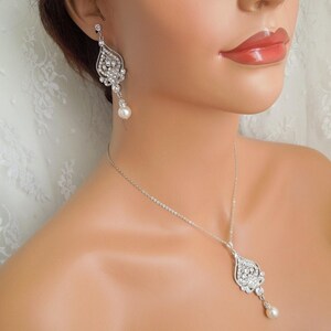 A mannequin wearing a silver-colored necklace and a matching earring, each with an ornate, filigree-style pendant encrusted with clear crystals and a single pearl drop.