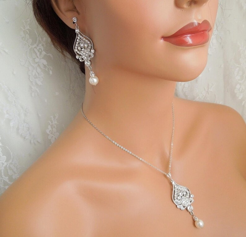 A mannequin wearing a silver-colored necklace and a matching earring, each with an ornate, filigree-style pendant encrusted with clear crystals and a single pearl drop.