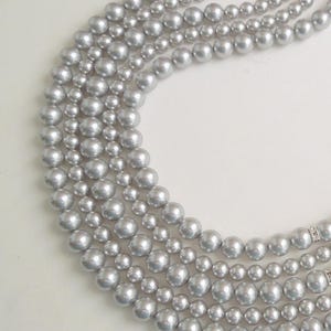 May include: A multi-strand necklace featuring four rows of round, metallic silver-toned beads. The beads are uniform in size within each strand, with the strands varying in length to create a layered effect. Small, clear crystal-encrusted spacers are visible on the strands.